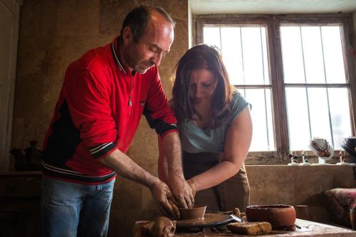 In a rustic room lit by two windows, a man in a red jacket and blue jeans guides the hands of the woman next to him, in a blue blouse and brown apron, both shaping a clay bowl on a spinning pottery wheel. Scattered around them are hunks of clay, finished pottery pieces, and a jewelry display in the windowsill.
