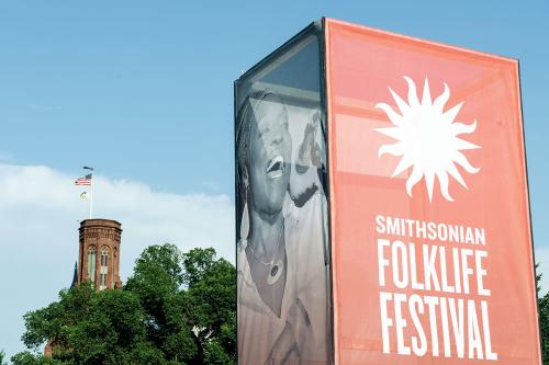 Tall event sign with the Smithsonian Folklife Festival logo in red on one side, photo of a woman singing on another, and the Smithsonian Castle seen in the background.