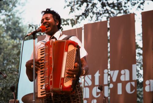 A man plays an accordion as he sings into a microphone on an outdoor stage.