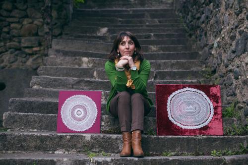 A woman sits in the middle of a stone stair case, smiling. To the left and right of her are placed examples of her needlework, made with white thread, mounted on purple velvet.