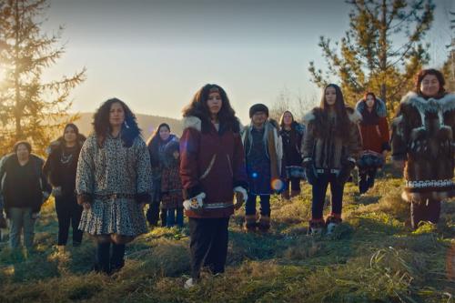 Group of Native women, all with straight black lines tattooed on their chins, stand pose on a hillside, the sun obscured by pine trees in the background.
