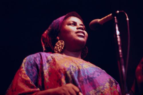 A woman with gold earrings and purple and orange matching blouse and headscarf speaks into a microphone on stage.