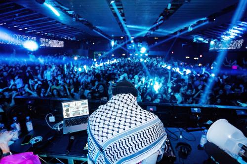 A person wearing a black beanie and white and black keffiyeh scarf faces away toward a packed crowd on a dancefloor, dimly light with dramatic blue rays of light.