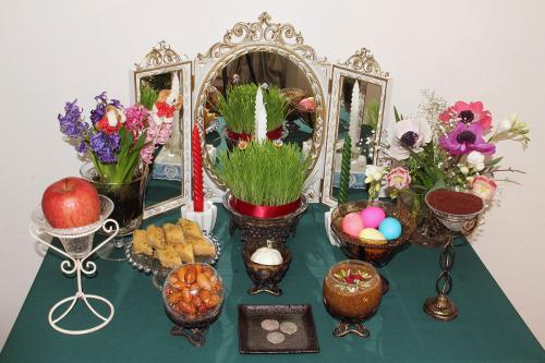 A turquoise table set with various objects: a mirror, potted grass, bouquets of flowers, an apple, dyed eggs, coins, candles, and other foods.