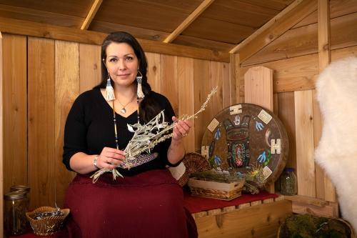 Seated in a small wooden room, a woman holds up dried sage, with Native art and baskets on the bench next to her.