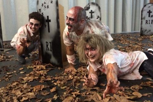 Three people act as blood-splattered zombies in a street filled with dead leaves and cardboard gravestones.
