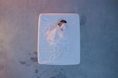 From above, a woman dressed in white, curled in fetal position, under a white sheet on a white mattress on a concrete floor.