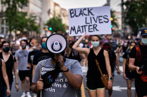 Countless people marching the street. In the foreground, a man with his face obstructed by a megaphone. Behind him, a woman holding up a sign reading BLACK LIVES MATTER. Behind them, out of focus, many people marching toward the camera, many in face masks.