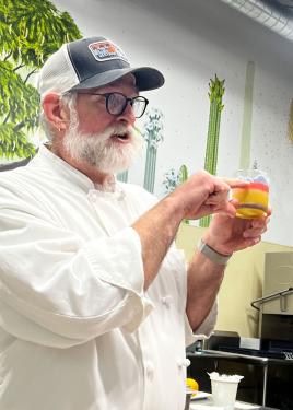 A man in a baseball cap and white chef's coat holds up a small clear cup with multicolored layers of curds: bright pink, white, yellow, and brown.