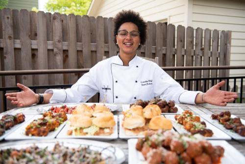 A Black woman in a white chef's coat opens her arms toward a table full of food.