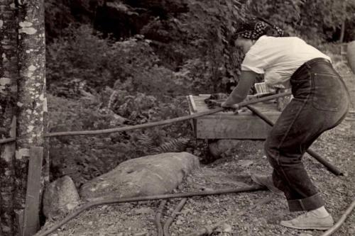 A woman in high-waisted jeans bends over in a clearing in the woods, pushing a long thin tool through a tree. The background is cluttered with other tools and sculpting materials.