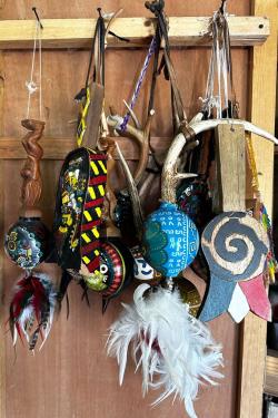 As assortment of wooden rattles and other musical instruments, carved, painted, and adorned with feathers, hang from a rack.