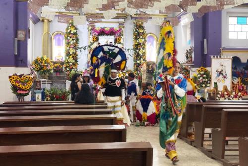 In a church with nearly empty pews, purple walls, floral decorations, and papel picado hanging from the ceiling, a few Feather Dancers prepare for their performance.