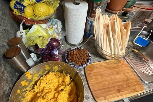 A kitchen counter set with a big bowl of yellow-orange masa, a bowl of dark brown shredded meat, a bowl of dried corn husks, and a cutting board.