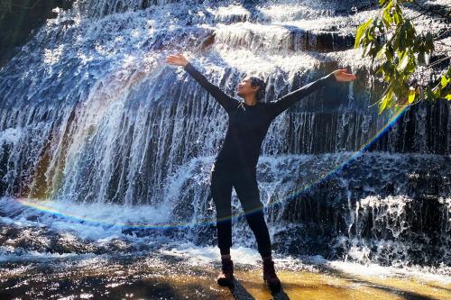 A woman sticks on a smooth rock surface in front of a wide waterfall. Its spray creates two arcs of rainbows that frame her, with arms outstretched.