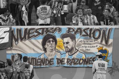 Banner in a soccer stadium with portraits of Maradona and Messi over the colors of the Argentina flag. The banner is in color, while the seated fans behind and film crew in front are in black and white.