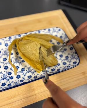 Close-up on the humita, unwrapped from its corn husk to expose a yellow-brown corn dough filling, as someone cuts into it with a fork and knife.