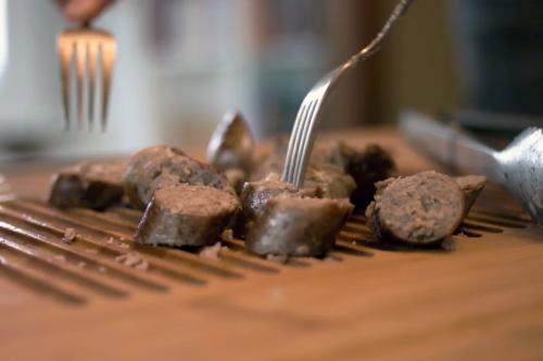Close-up on two forks reaching for slices of sausage on a cutting board. 