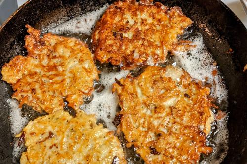 Four golden brown, shredded potato pancakes frying in a cast iron pan.