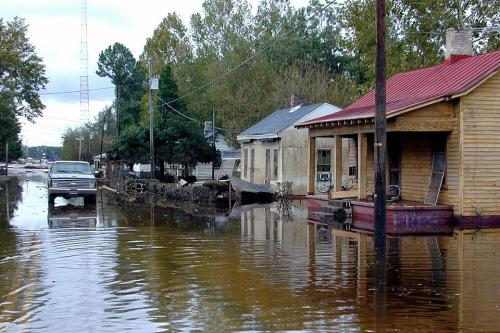 A flooded street in a town, with muddy water up to the edges of single-story wooden homes. A pickup truck drives down the road regardless.