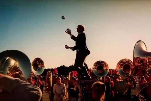 At dusk, silhouetted figure of a man juggling, surrounded by the glinting bells of sousaphones and marching band members in uniform.