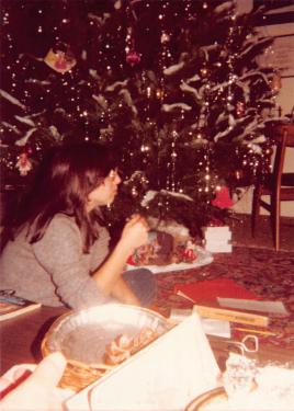 A teen girl with long, straight brown hair lies on a floor at the foot of a decorated Christmas tree, propped up on her elbows. On some, but not all, of the branches is white cotton fluff resembling snow.