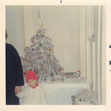A young child wearing a white coat and red beanie holds the hand of a person wearing a dark coat, their head out of frame, standing in front of a tinsel-adorned Christmas tree.