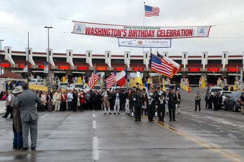 People dressed in military uniform and formal wear cluster at the start of a parade route on a wide road. In the background, an American flag flying above a banner that reads 'Bienvenidos Washington's Birthday Celebration,' and a solid line of inspection booths at an international border.