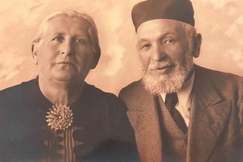 Portrait of a woman with straight white hair pulled back, a dark blouse, and flower broach, and a bearded man in a dark suit and cap. Sepia-toned black-and-white photo.