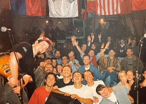 A man with a dark goatee and a red ball cap, holding an electric guitar, poses in front of a cheerful audience in a small music venue. Above them hang several national flags.