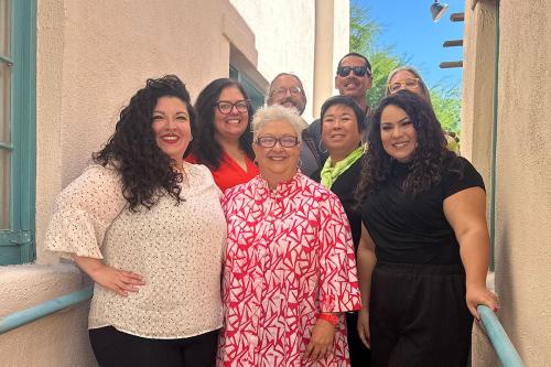 Eight adults pose in an outdoor corridor, all smiling broadly.