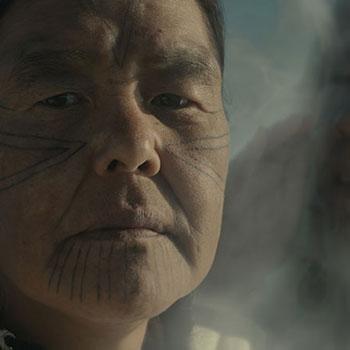 Film still of a closeup of a person's face, with tattooed lines on their cheeks and chin, and a stern look.