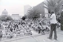 A man stands on an outdoor stage, speaking through a microphone to a large seated crowd, with a domed building in the distance. Black-and-white photo.