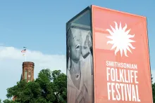 Tall event sign with the Smithsonian Folklife Festival logo in red on one side, photo of a woman singing on another, and the Smithsonian Castle seen in the background.