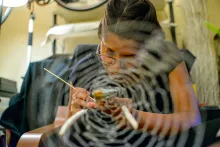 A woman leans over the work in her hands, holding a paintbrush or other long tool. She is partly obscured by a spider web-like tapestry in front of her.