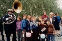 A man dressed in a black suit blows into a sousaphone, posing with a group of young kids.