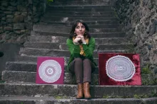 A woman sits in the middle of a stone stair case, smiling. To the left and right of her are placed examples of her needlework, made with white thread, mounted on purple velvet.
