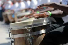Close-up on a pair of Black hands playing a drums. In the blurry background, a whole line of seated drummers.