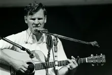 A white man plays acoustic guitar on stage, with two microphones and a harmonica in front of him and a harmonica holster around his neck. Black-and-white photo.