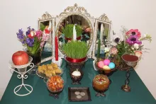 A turquoise table set with various objects: a mirror, potted grass, bouquets of flowers, an apple, dyed eggs, coins, candles, and other foods.