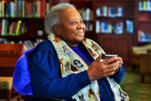 An older woman sits and smiles in a library room, holding a harmonica. Her scarf is printed with cartoon dogs.