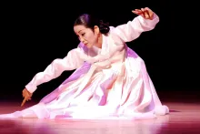 A woman in a long-sleeved pink gown crouches on a stage in a dance, with a spotlight on her.