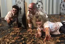 Three people act as blood-splattered zombies in a street filled with dead leaves and cardboard gravestones.