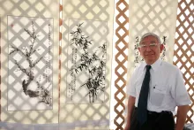 A elder man with short gray hair, glasses, and a blue tie with white button-up shirt poses besides hanging vertical paintings, depicting plum blossoms and bamboo with Chinese characters.