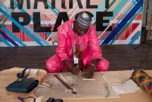A man in a pink suit sits on the ground, learning over a workspace. In front of him is a piece of leather and leather tools, which he uses as he works on the leather making a bag.