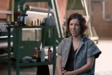 A woman with short wavy hair sites in a workroom, speaking to a camera out of frame. Behind her is work bench with tools, a ream of paper, and other supplies.