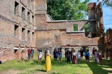 Rows of teens stand in formation, socially distanced, with a woman in a bright yellow dress standing in front, conducting them. They are outdoors, surrounded by the remnants of a dilapidated, roofless brick building.