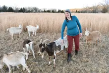 A woman pets one of six goats on a farm in winter, with bare trees and a field of golden grain behind her.