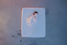 From above, a woman dressed in white, curled in fetal position, under a white sheet on a white mattress on a concrete floor.
