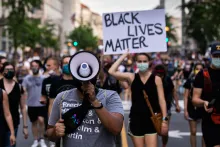 Countless people marching the street. In the foreground, a man with his face obstructed by a megaphone. Behind him, a woman holding up a sign reading BLACK LIVES MATTER. Behind them, out of focus, many people marching toward the camera, many in face masks.